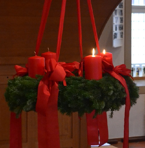Das heutige Bild zeigt einen großen Adventskranz, der an 4 roten Bändern hängt. Auf dem Kranz sind 4 rote Kerzen befestigt. Zwei Kerzen brennen. Wir sagen euch an den lieben Advent! 