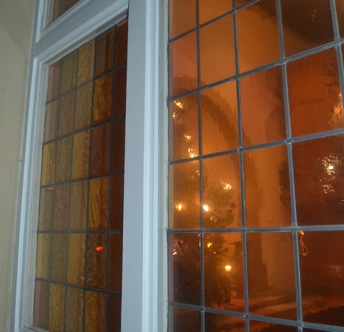 Das Bild zeigt einen Teil eines zweiflügeligen geschlossenen Fensters. Der Fensterrahmen ist weiß lackiert, die Fensterfläche besteht aus kleinen bleiverglasten Scheiben. Durchs Fenster scheint warmgelbes Licht von einzelnen Lampen, die wie Sterne im Raum schweben. Wie im Nebel erkennt man die Umrisse einer Tanne. Es könnte ein Weihnachtsbaum sein!