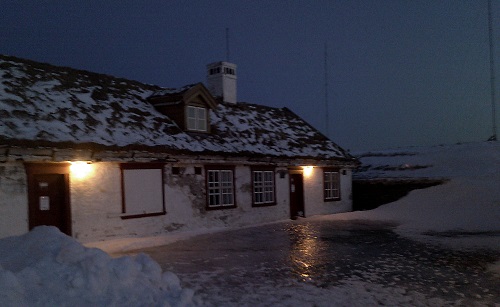 Auf unserem Bild ist es Winter geworden: Schneegrauer Winterhimmel, Schneeberge und eine riesige, glänzende Eisfläche vor einem niedrigen Haus. Am Haus brennen zwei Außenleuchten, deren Licht sich auf der dunkeln Eisfläche spiegelt.