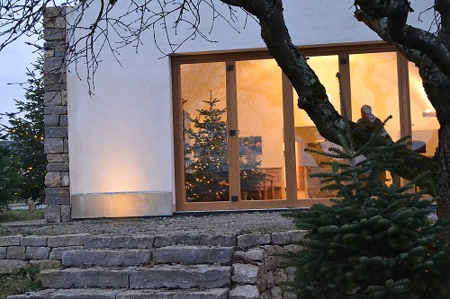Winterabendstimmung: Eine Treppe aus groben Natursteinen führt auf eine Terrasse. Die Scheiben der Terrassentür  leuchten in warmgelbem Licht. Im Zimmer steht ein schön geschmückter Weihnachtslichterbaum.