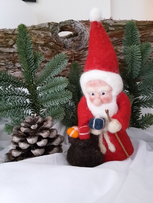 Das heutige Kalenderfensterchen zeigt einen kleinen Weihnachtsmann aus Filz. Der kleine Kerl trägt einen roten Mantel und eine rote Kapuze. Sein Gesicht mit der Knollennase und den frostgeröteten Bäcklein ist eingerahmt von einem schneeweißen Bart und weißen Haaren. Den Sack hat der Kleine vom Rücken genommen und offen neben sich in den Schnee gestellt. Im Sack sieht man ein orangenes und ein rotes Päckle.  Ein blaues Päckle hat der Weihnachtsmann aus dem Sack genommen und hält es Dir entgegen. Frohe Weihnachten!